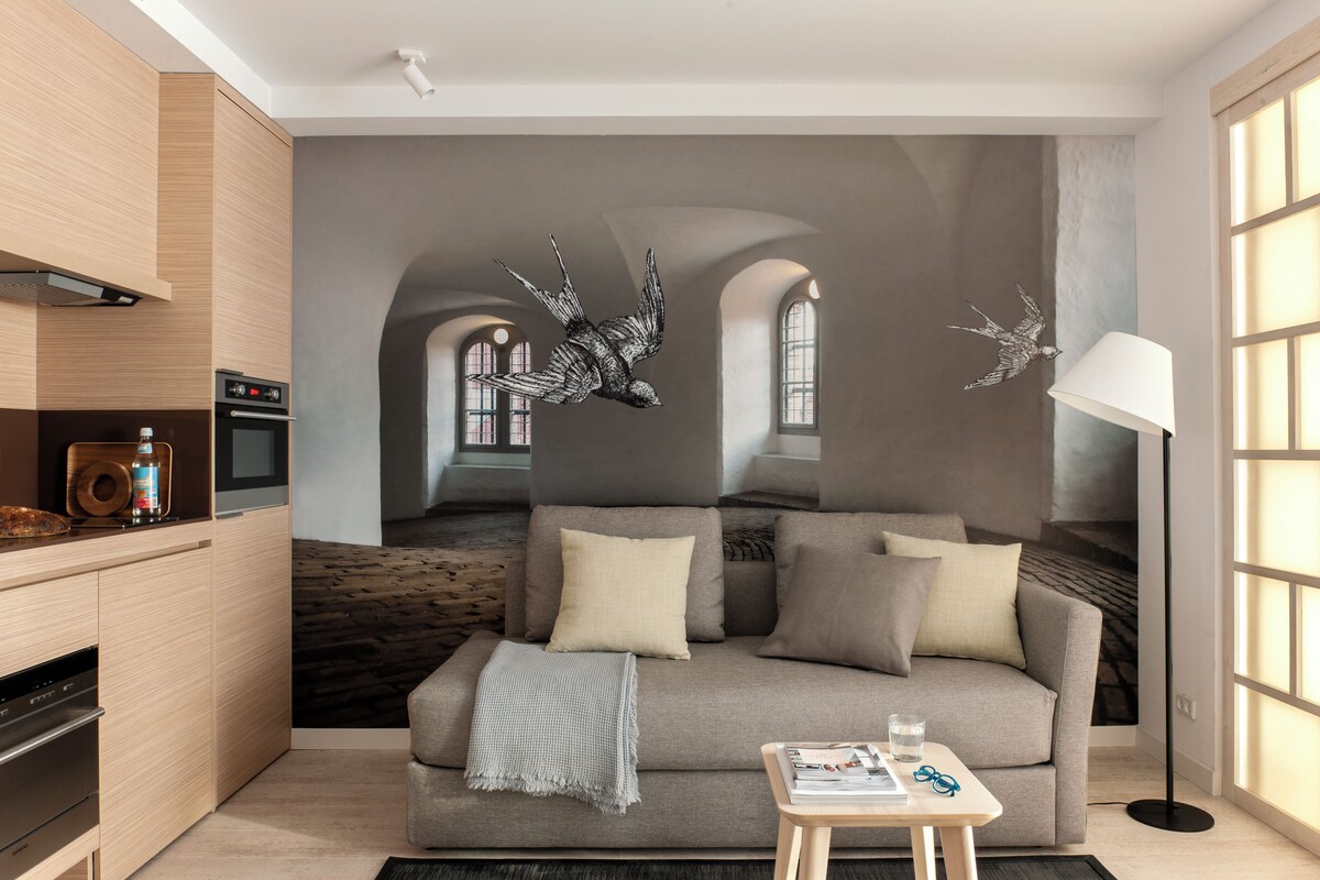 Ein modernes Wohnzimmer mit einem grauen Sofa, einem Holztisch und einer Stehlampe. An der Wand befindet sich ein Wandbild von einem Vogel, und es gibt eine Küchenzeile mit Herd und Backofen.