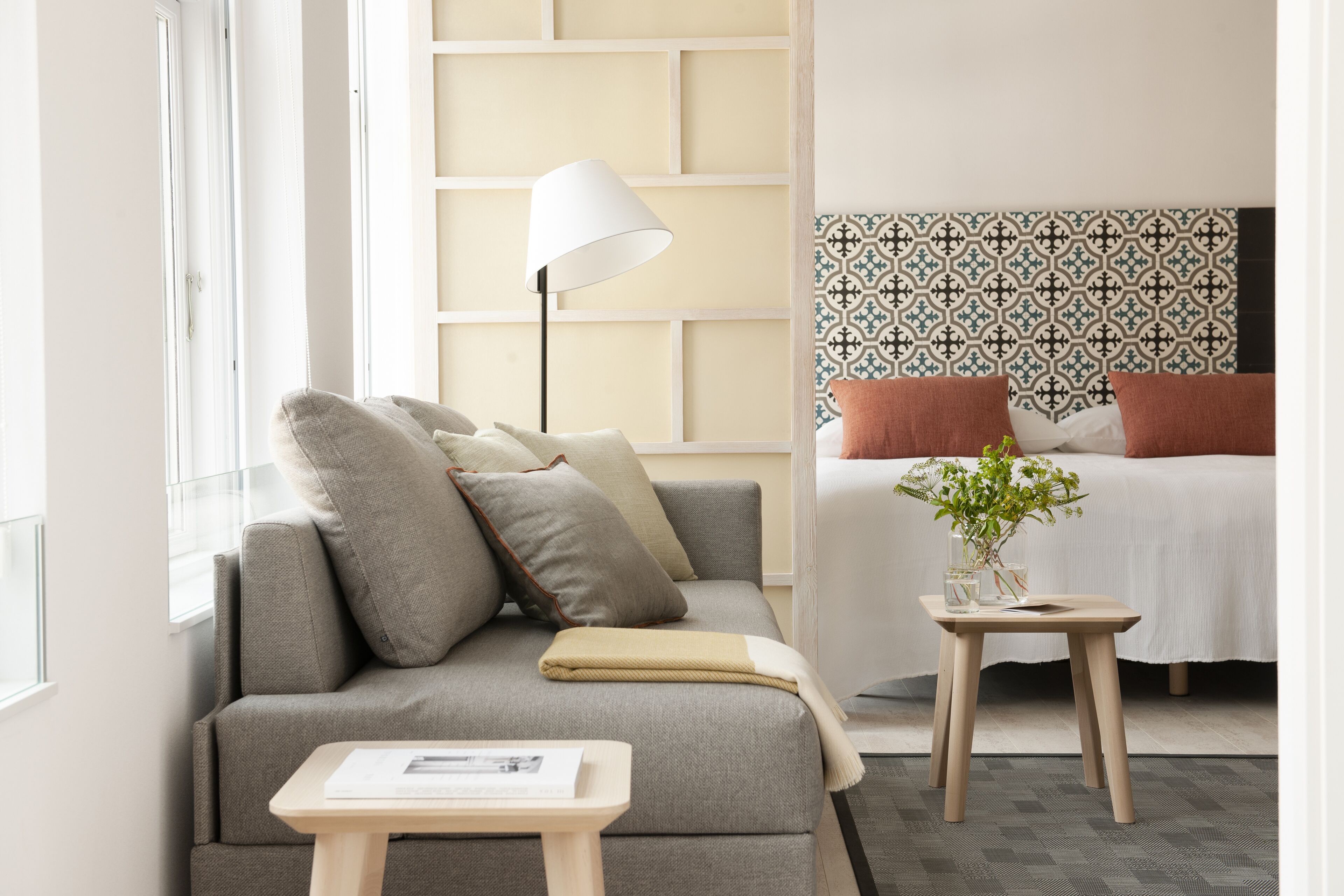 Una habitación de hotel moderna con sofá, mesita de noche, lámpara, cama con cabecero estampado y una planta pequeña sobre la mesita.
