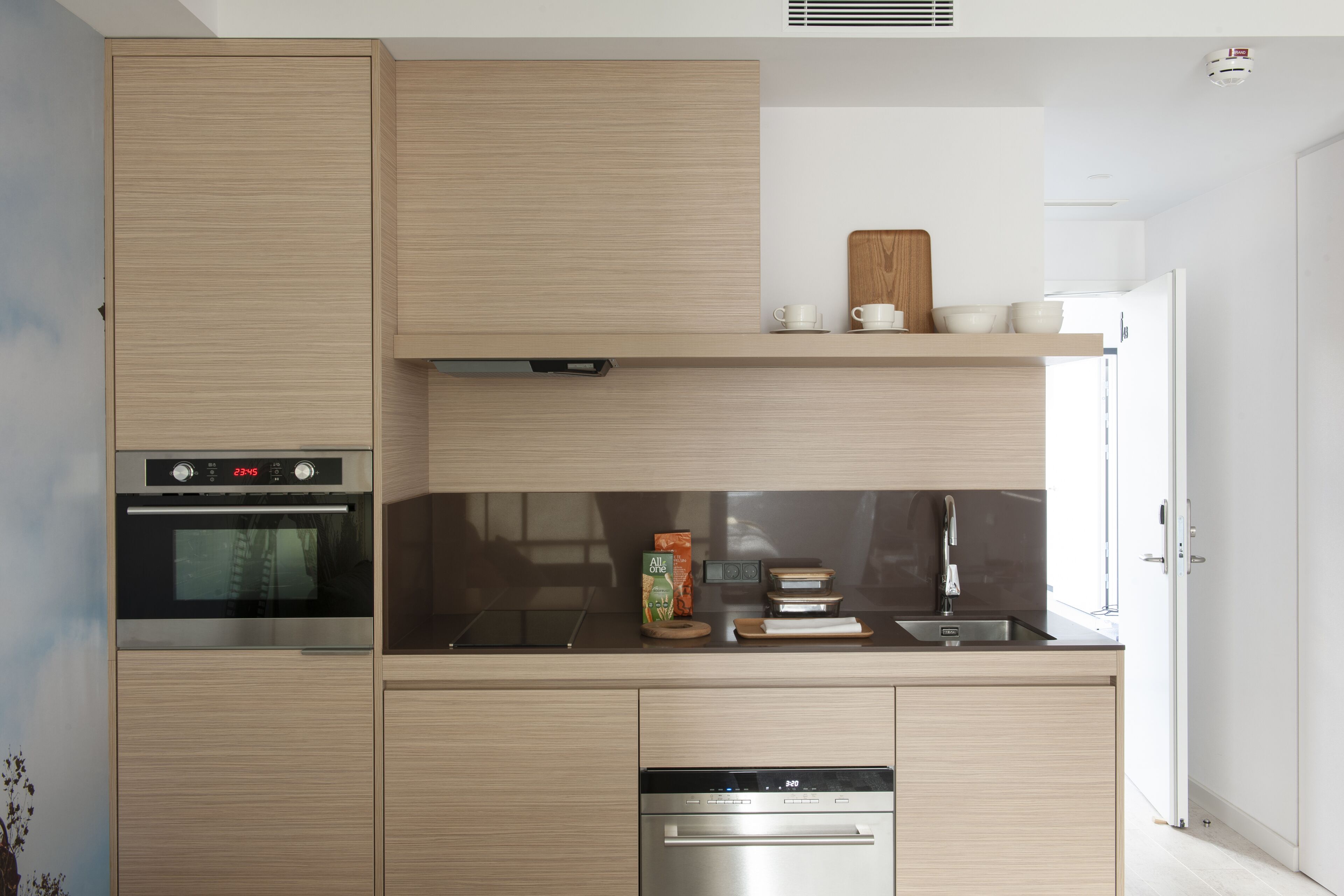 Una cocina moderna con armarios de madera, horno integrado, lavavajillas y una pequeña encimera con cafetera y tazones.