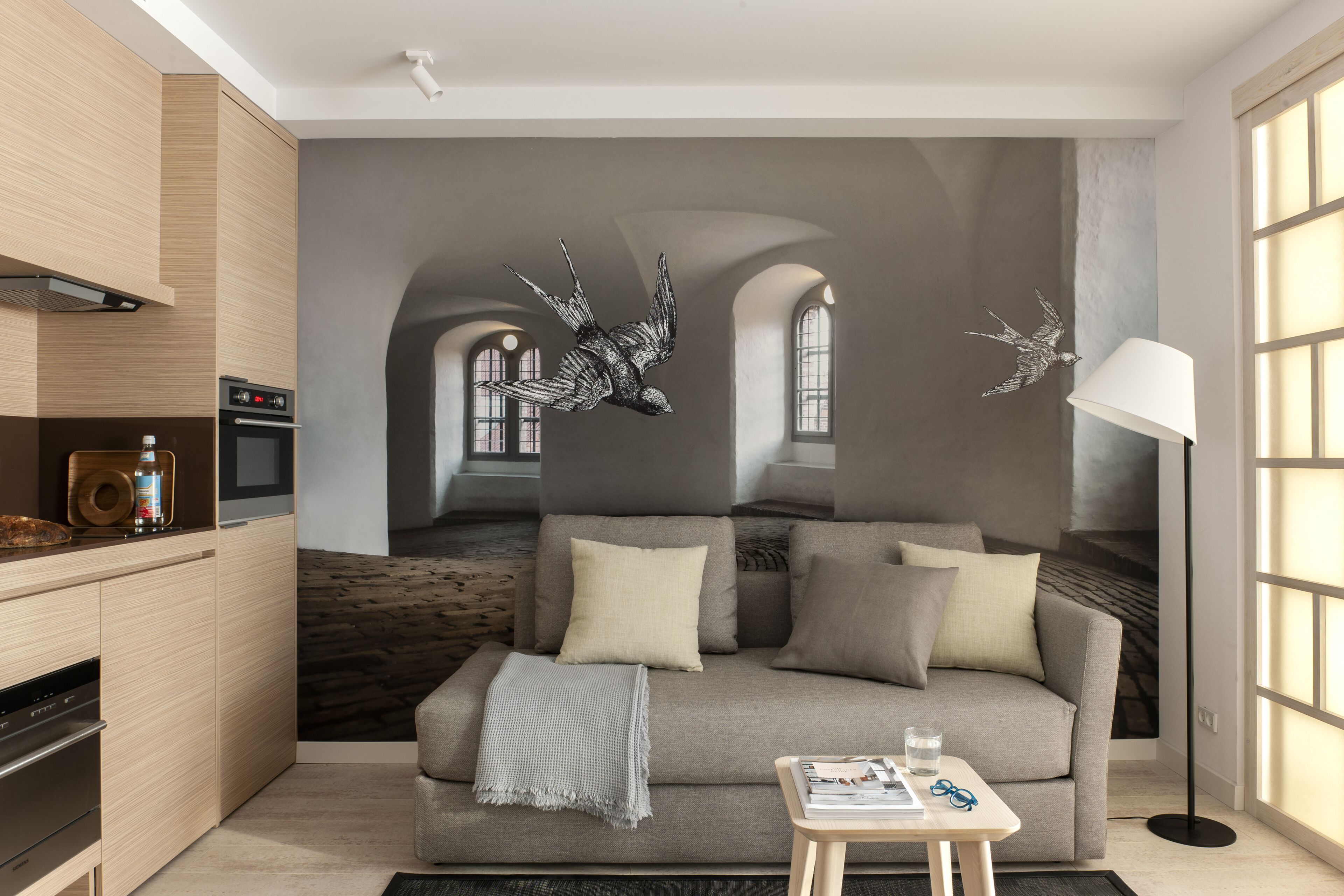 A modern living room with a grey sofa, a coffee table, and a lamp. There is a kitchen area with a stove and oven.