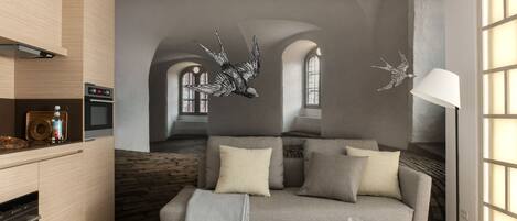 A modern living room with a grey sofa, a coffee table, and a lamp. There is a kitchen area with a stove and oven.