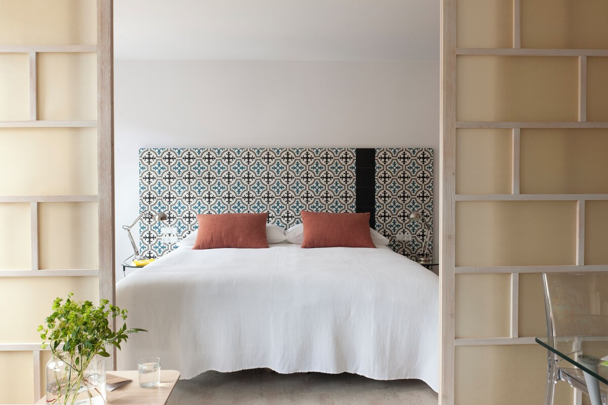 Ein modernes Schlafzimmer mit einem gemusterten Kopfteil, einem Bett mit weißer Bettwäsche und zwei Kissen, einem Nachttisch mit Lampe sowie einem Glastisch mit einer Vase und einem Glas Wasser.