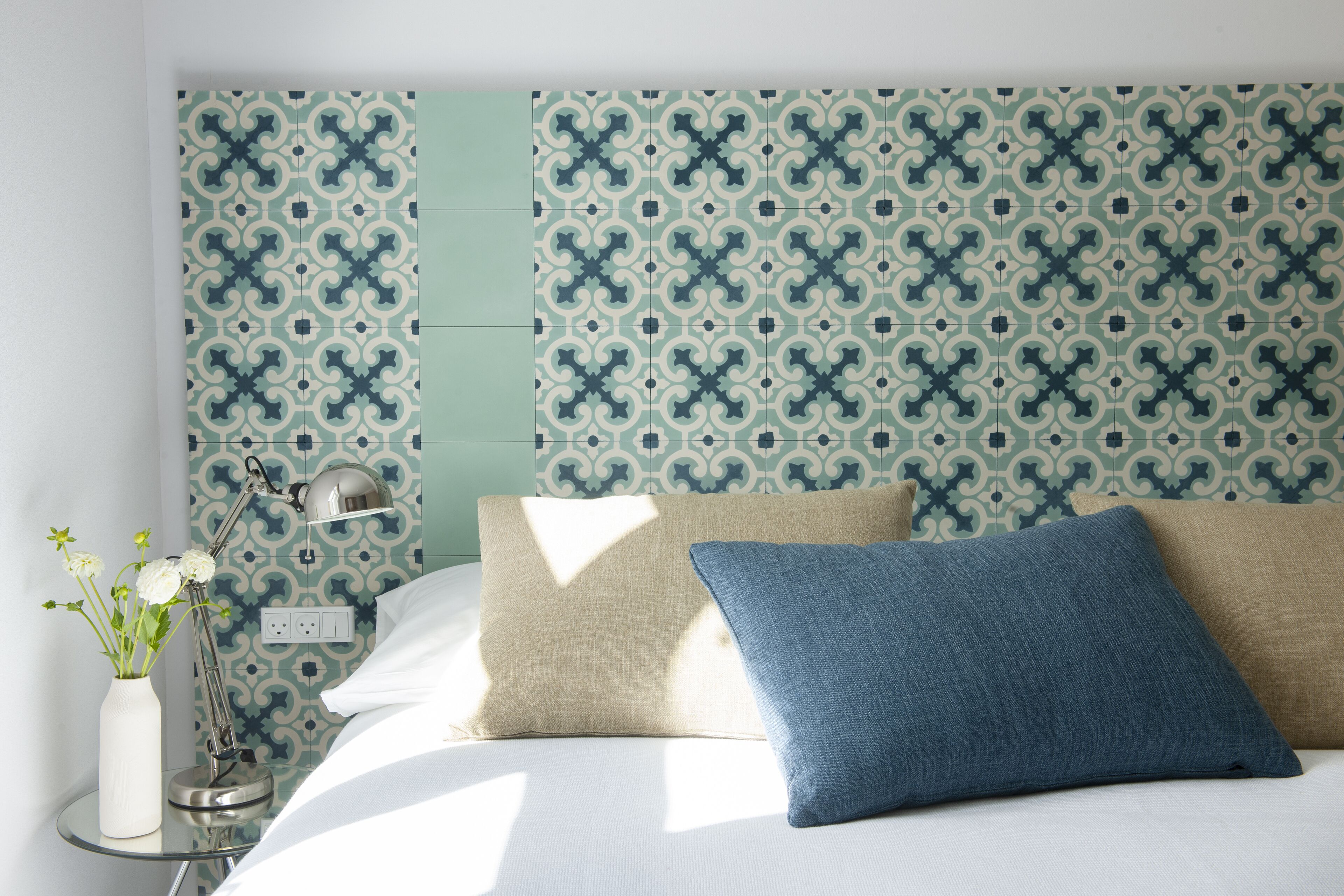Cama bem arrumada com duas almofadas, abajur e um vaso com flores em uma mesa de cabeceira.
