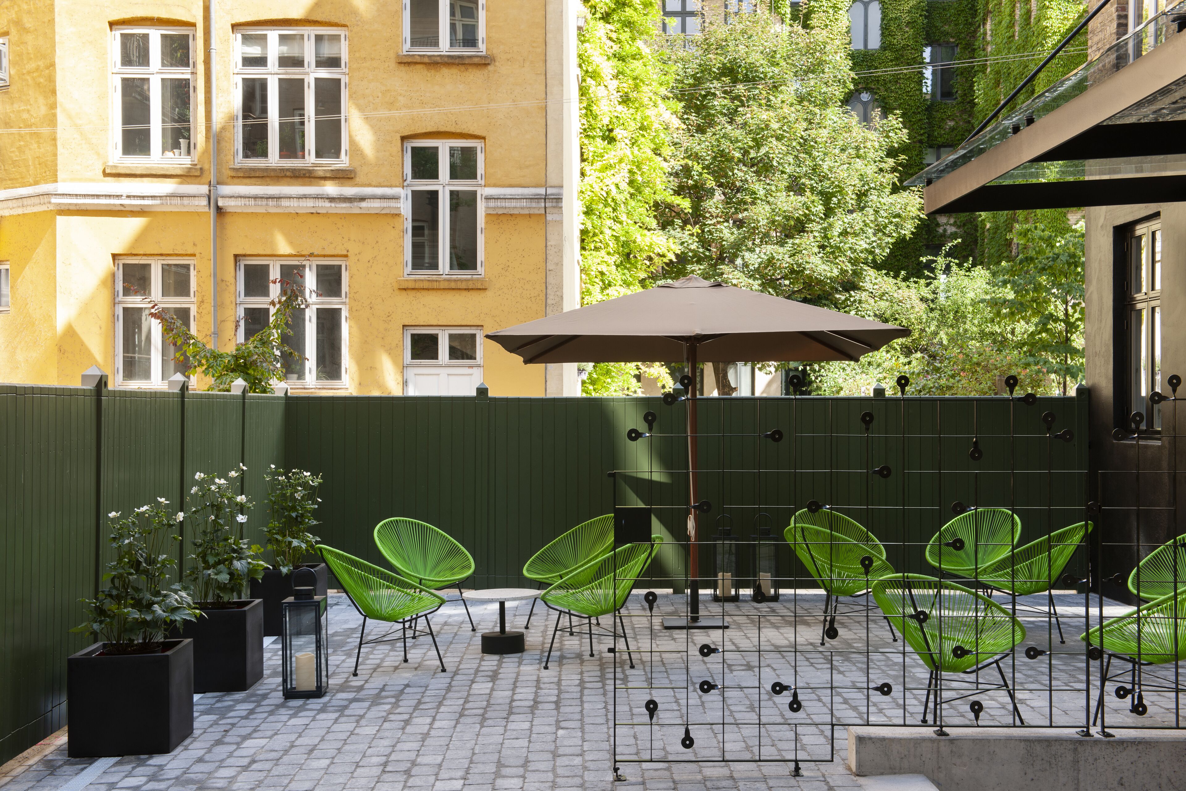 Un área de estar al aire libre con sillas verdes y una mesa bajo una sombrilla, rodeada por una cerca verde y plantas en macetas.