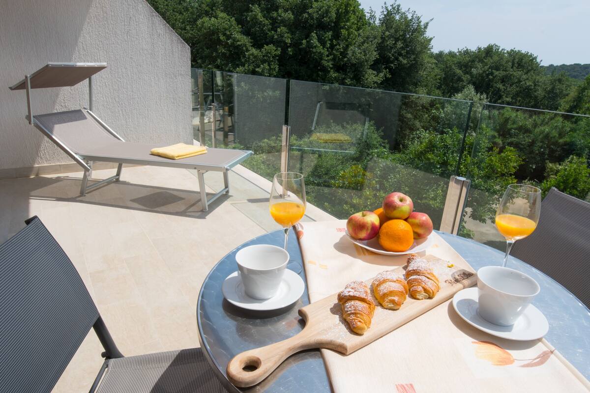 A dining table with a breakfast setup including croissants, apples, oranges, and juice glasses, overlooking a lush green landscape.