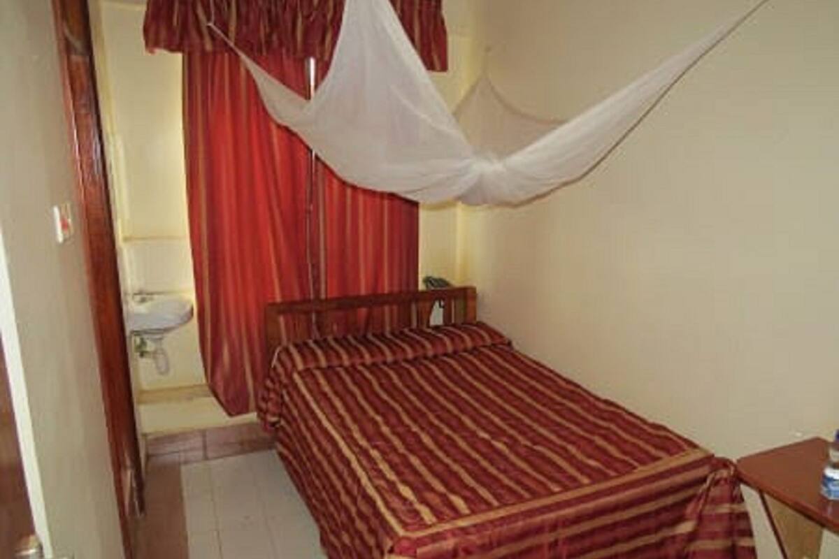 Ein Einzelbett mit gestreifter Bettdecke, ein Nachttisch mit einer Flasche und ein Vorhang in einem Zimmer mit Fliesenboden und einem sichtbaren Waschbecken.
