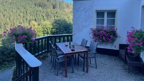 A terrace with a table and chairs, surrounded by flower boxes with pink flowers, set against a forested hillside. - Haus Onel 2 (Nordenau)