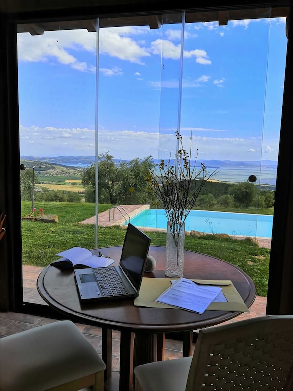 Mesa redonda com um laptop, papéis e um vaso com galhos. Uma piscina é visível ao fundo.