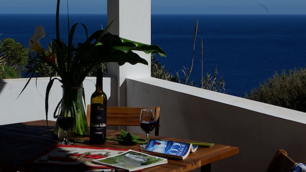 Une table avec une bouteille de vin, deux verres, un livre et un vase de fleurs, le tout placé sur un balcon donnant sur la mer.