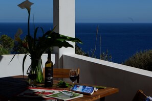 Une table avec une bouteille de vin, deux verres, un livre et un vase de fleurs, le tout placé sur un balcon donnant sur la mer.