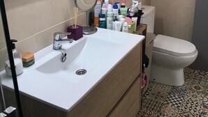 Et badeværelse med en hvid vask, spejl og toilet. Der er toiletartikler på vasken og et vindue over toilettet.