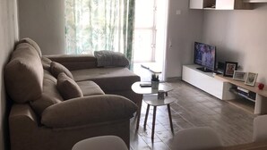 En moderne stue med en modulsofa, et lille rundt bord, et fjernsyn på et hvidt skab og en geometrisk hængende lampe.