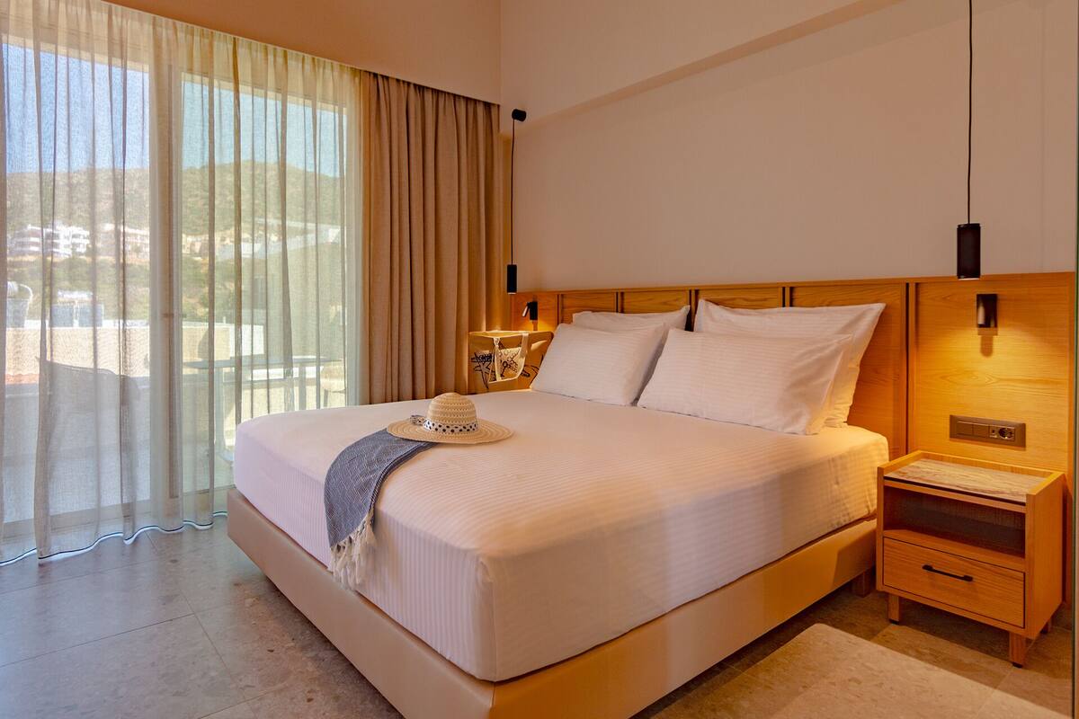 Une chambre d’hôtel avec un grand lit, une table de chevet, une lampe et une vue sur l’extérieur à travers des rideaux transparents.