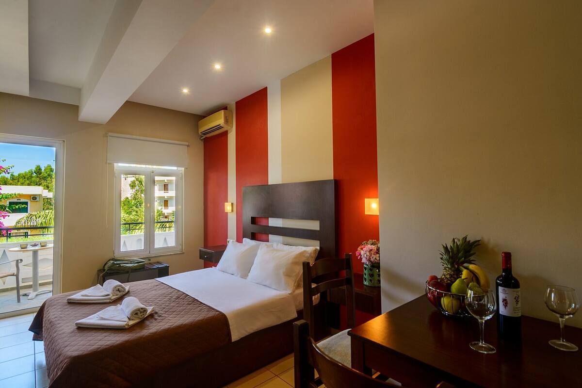 Une chambre d’hôtel avec un lit, une table à manger avec une bouteille de vin et un verre, un panier de fruits et une vue sur l’extérieur.