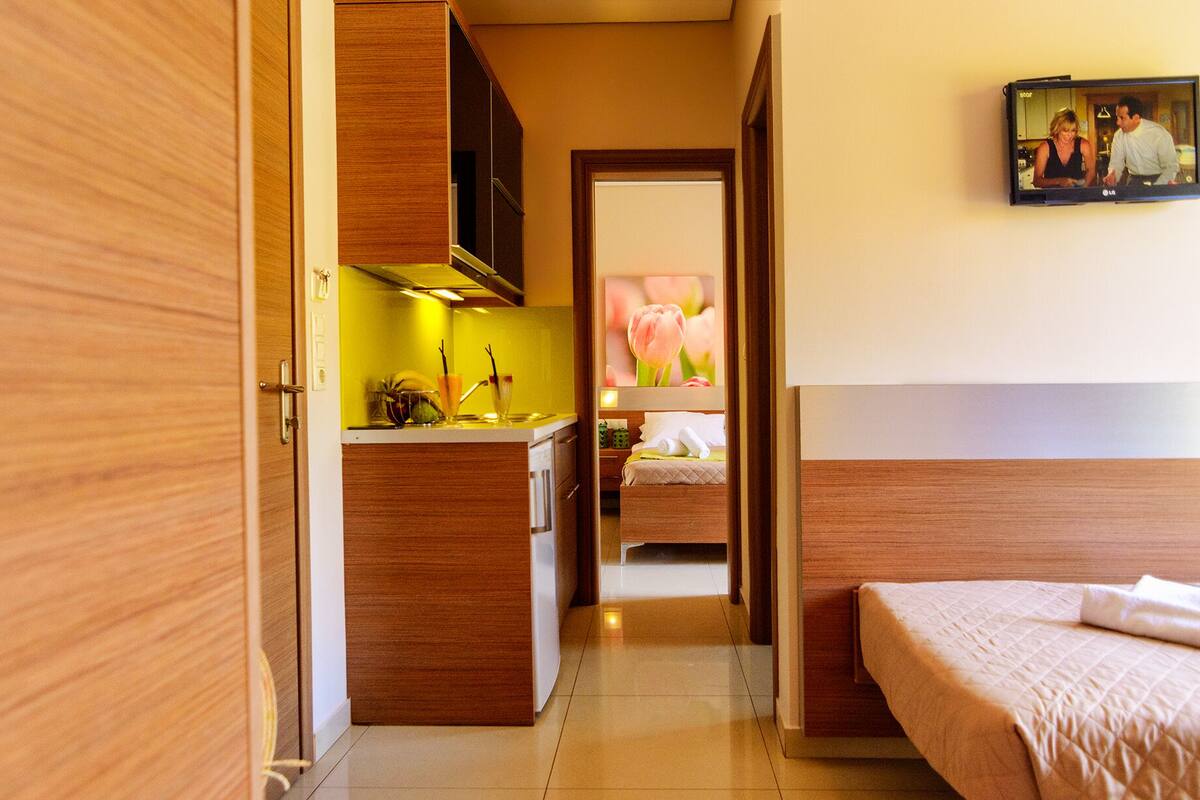 Une chambre d’hôtel avec une armoire en bois, une kitchenette dotée d’un plan de travail jaune, un lit recouvert d’une courtepointe matelassée et un téléviseur diffusant une image d’un couple.