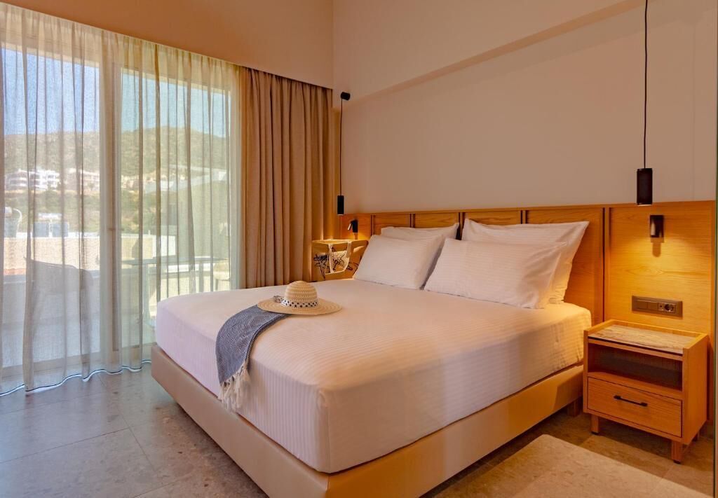 Une chambre d’hôtel avec un grand lit, une table de chevet, une lampe et une vue sur l’extérieur à travers des rideaux transparents.
