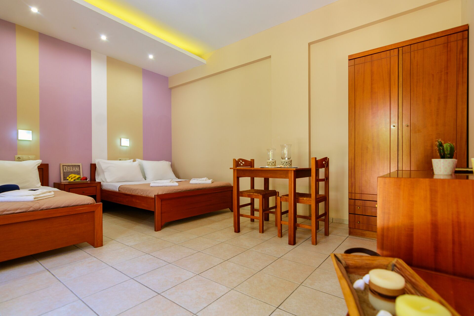 A hotel room with two beds, a wooden wardrobe, a small table with chairs, and a tray with candles.
