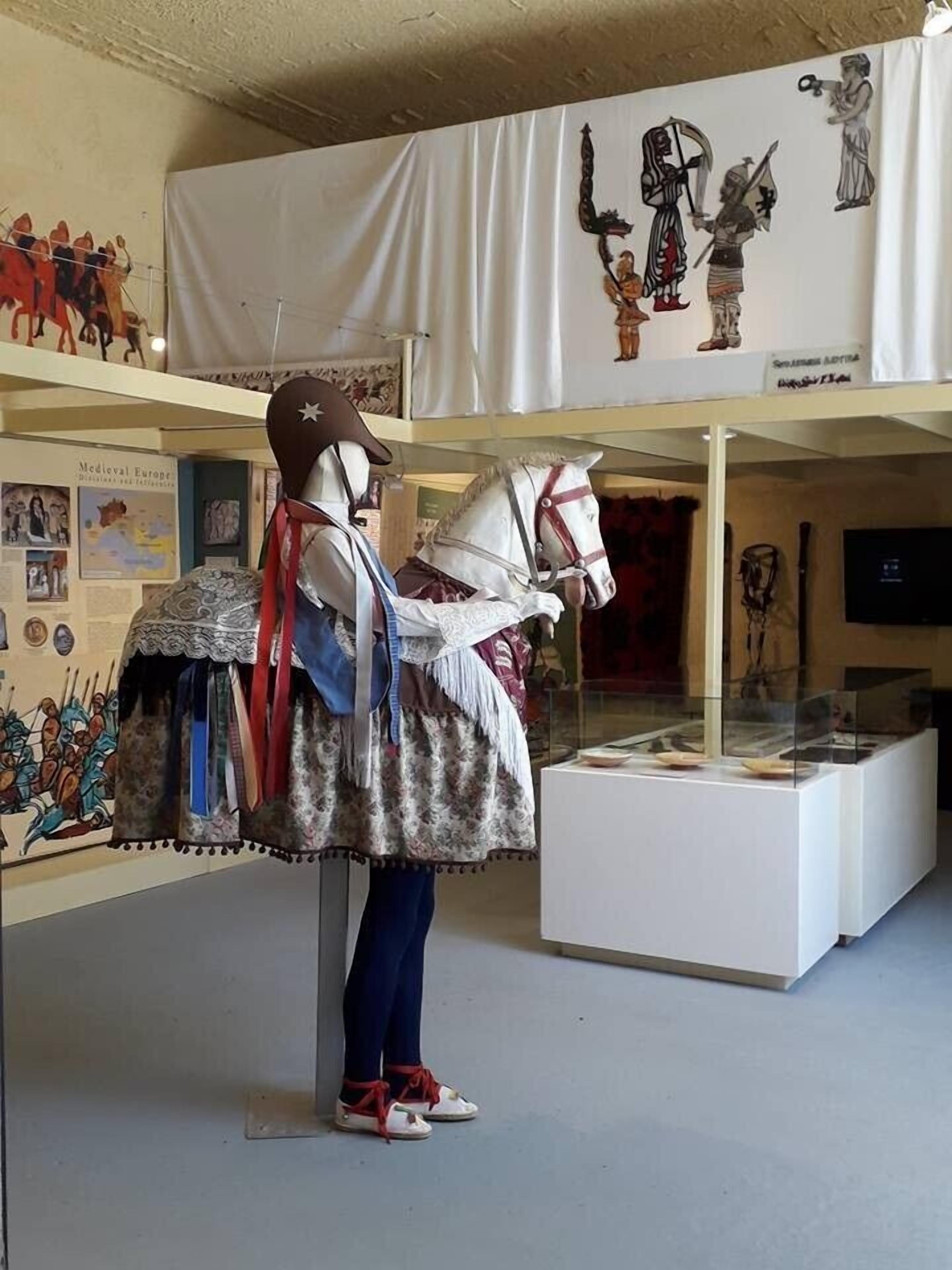 Een etalagemodel gekleed in middeleeuwse kleding, staand in een museumexpositie met historische voorwerpen en een banner waarop middeleeuwse figuren afgebeeld zijn.
