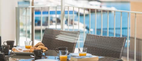 A breakfast table with pastries, juice, and a book on a balcony overlooking a marina.