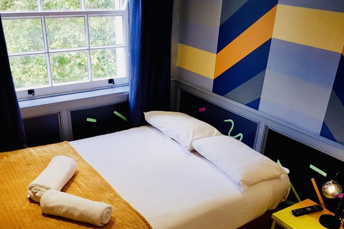 Hotellihuone, jossa on sänky, ikkuna verhoilla, yöpöytä lampun kanssa ja seinä, jossa on geometrisia kuvioita.