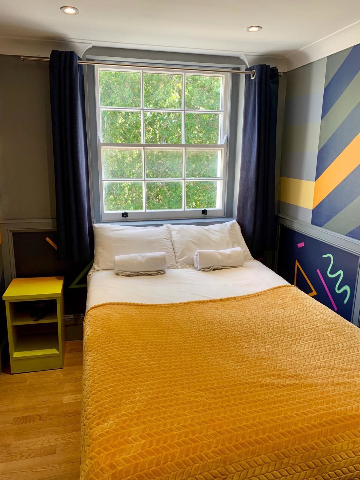 A bedroom with a large window, a bed with white linens, a yellow quilt, and a green nightstand.