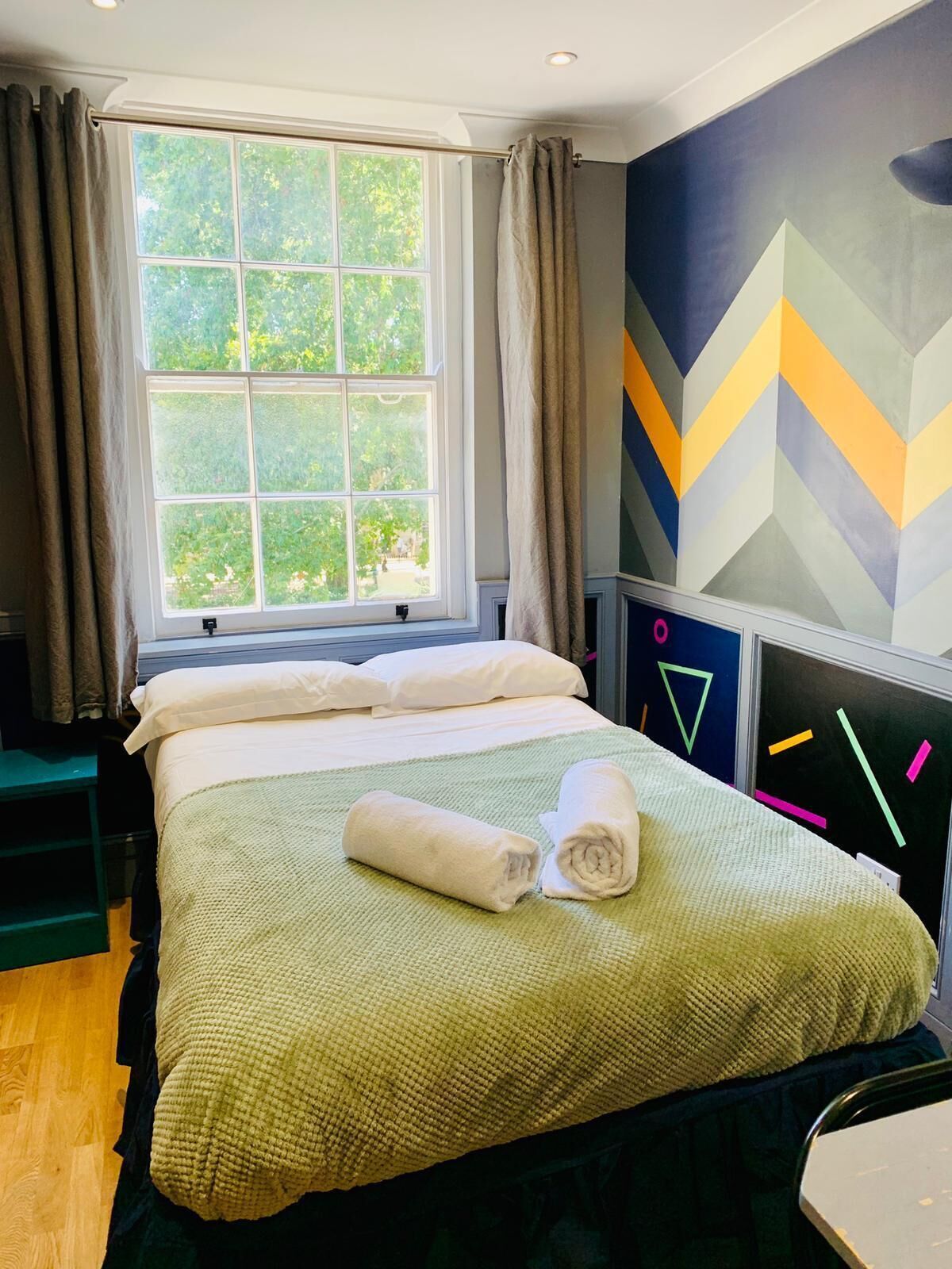 Een hotelkamer met een bed, een raam met gordijnen, een geometrisch wandontwerp en uitzicht op groen.