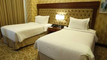 A hotel room with two beds, a wooden headboard, a bedside table with a lamp, and a patterned carpet.