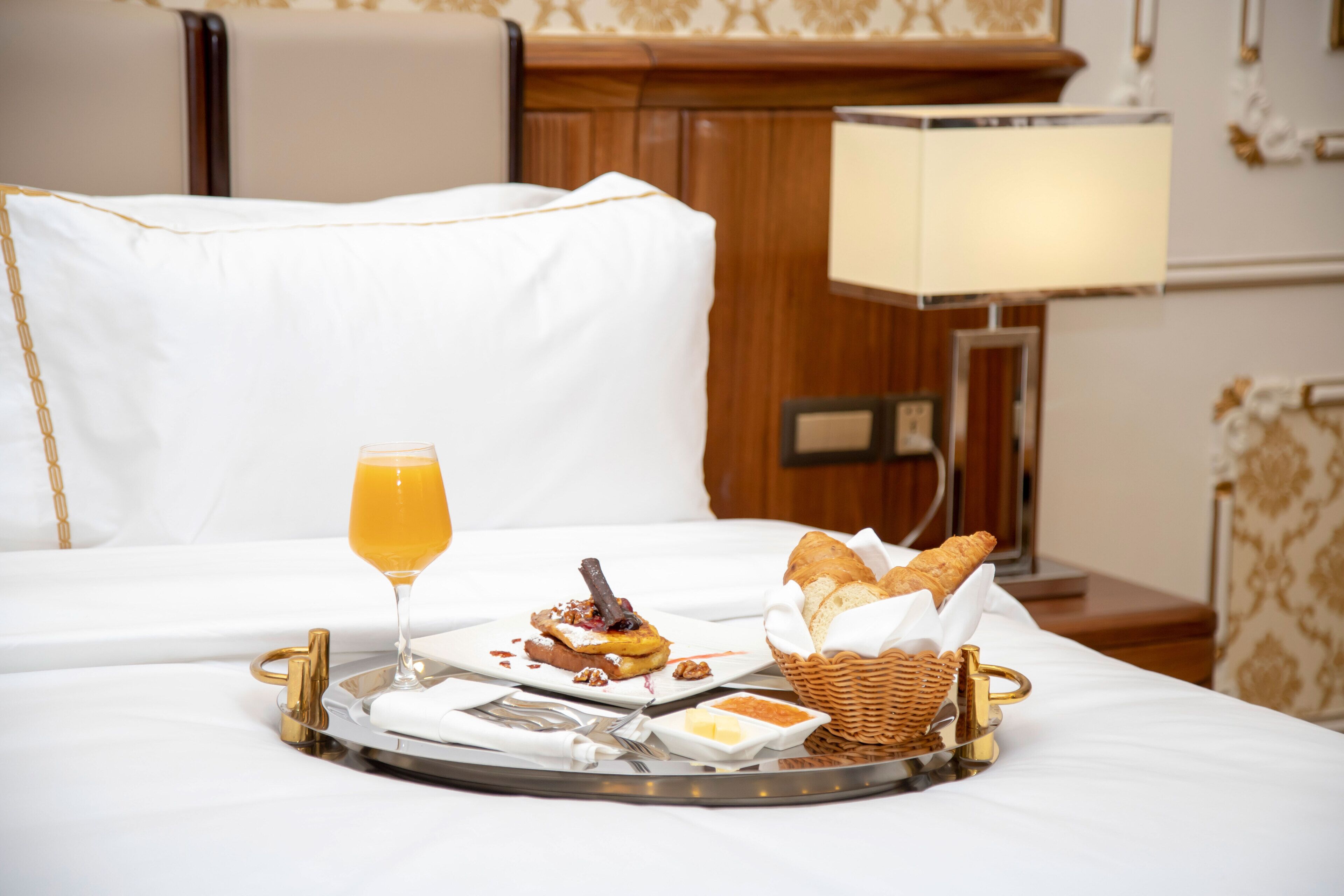 A breakfast tray with toast, a glass of orange juice, and a basket of bread on a bed in a hotel room.