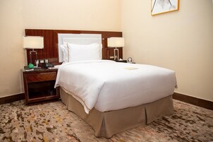 Quarto de hotel com uma cama grande, duas mesas de cabeceira com luminárias, uma cabeceira de madeira e um tapete estampado.