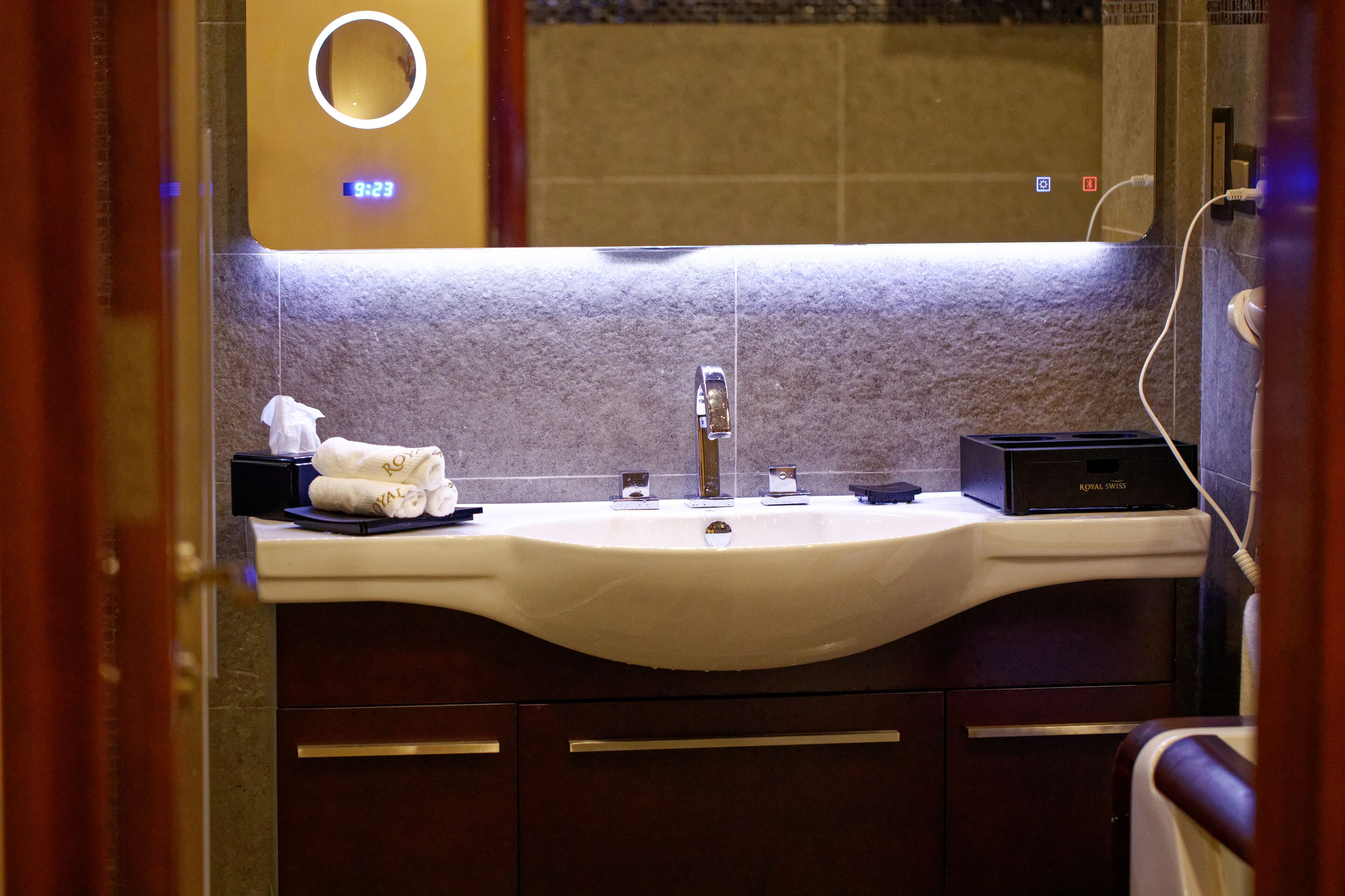 Une salle de bain avec un lavabo blanc, un miroir et une horloge numérique affichant 9h23.