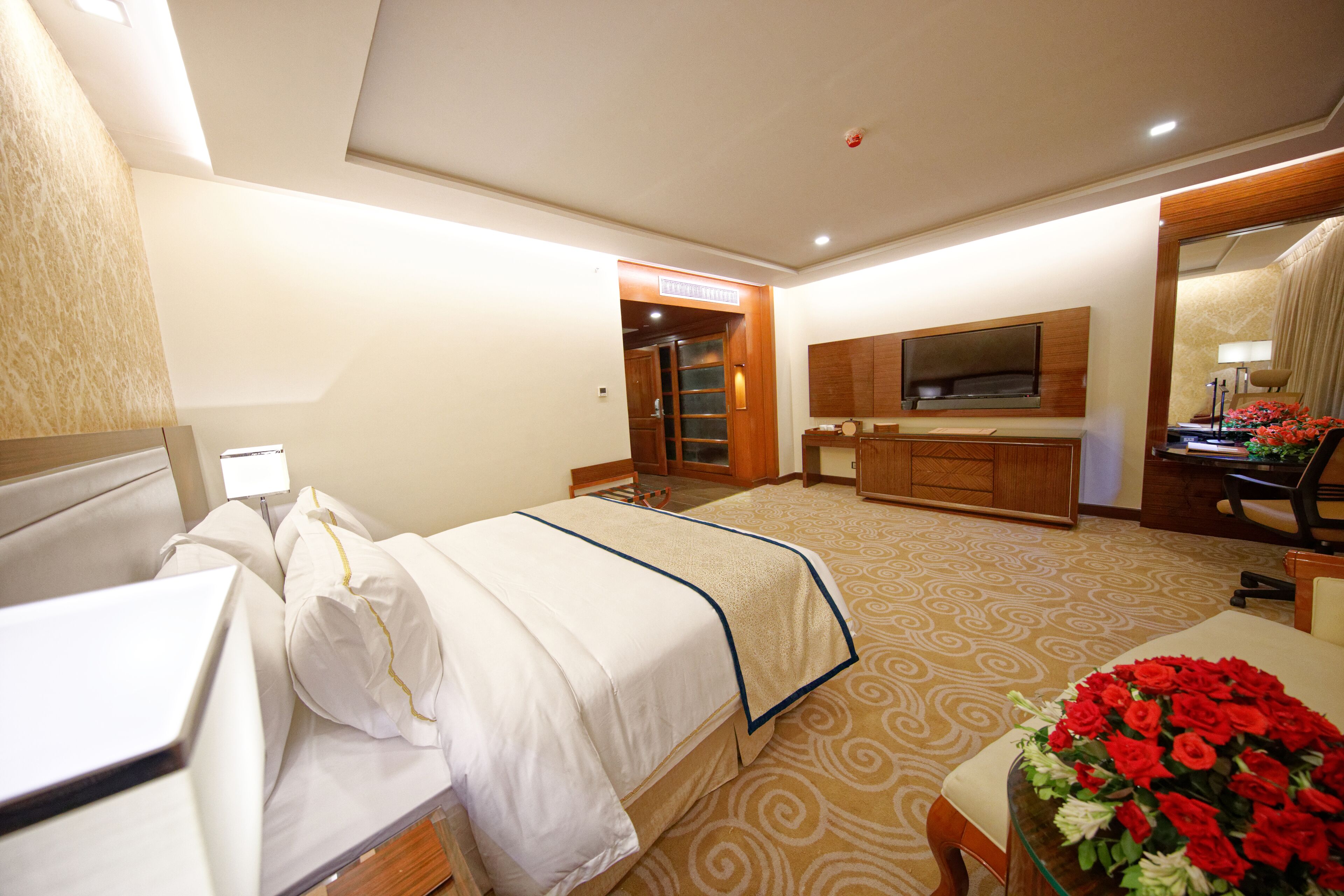 A hotel room with a bed, a desk, a TV mounted on the wall, and a floral arrangement on the table.