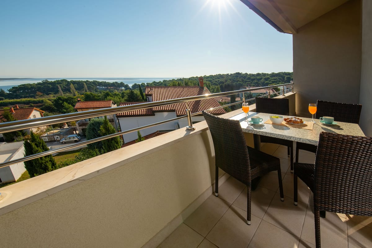 A balcony with a table set for two, overlooking a scenic view of a coastal area with houses and trees.