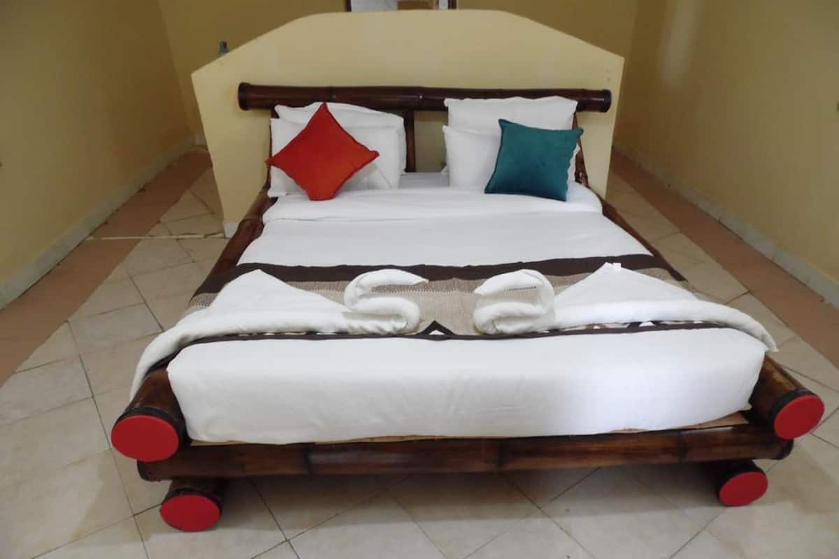 Un lit double avec un cadre en bois et des roulettes rouges, une literie blanche et deux oreillers (un rouge, un turquoise).