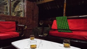 A cozy room with a stone wall, a wooden beam, and a red sofa with a green checkered throw. There are two glasses of a golden liquid on a white table.