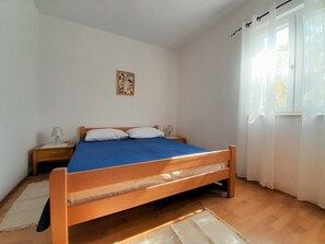 A bedroom with a wooden bed, white walls, a small bedside table with a lamp, and a window with sheer curtains. - Apartment Tihana with 2 bedrooms and garden view, family-friendly (Krk)