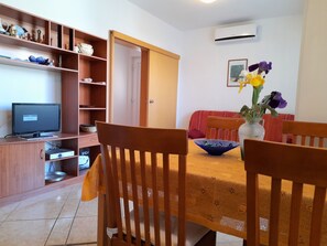A dining area with a wooden table, chairs, a TV, and a shelf with decorative items. - Apartment Tihana with 2 bedrooms and garden view, family-friendly (Krk)