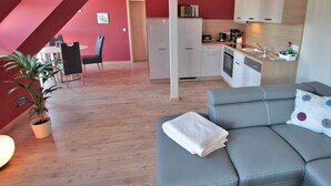 A modern living room with a grey sofa, a small table, and a potted plant. A kitchen with white cabinets and a sink is visible in the background. - Apartment for a perfect holiday with the family (Greifswald)