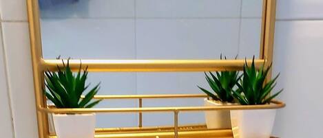 A bathroom with a gold-framed mirror, two white planters with green plants, and a towel hanging on the wall.