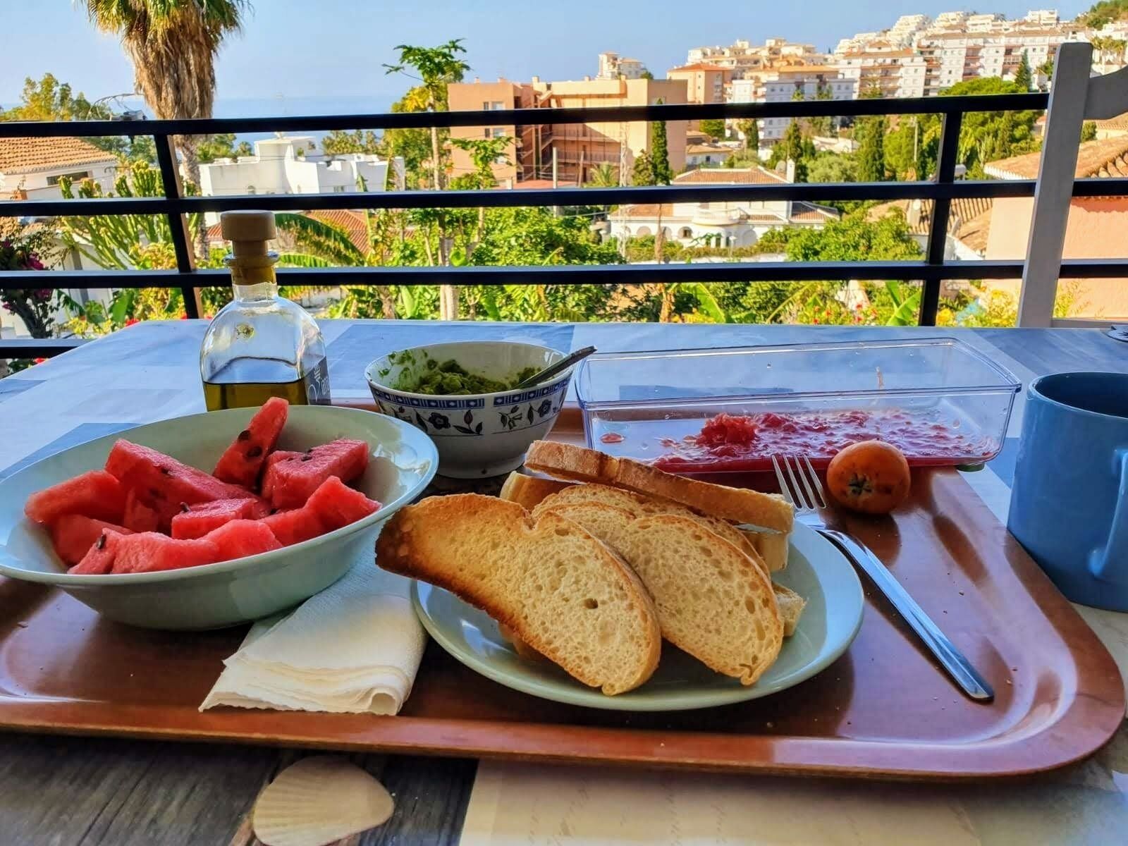 Una mesa con una bandeja que contiene sandía, pan y un tazón de guacamole, con vistas a edificios y follaje al fondo.