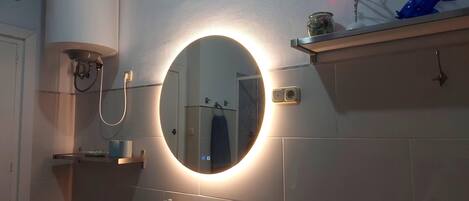 Une salle de bain avec un miroir rond, un lavabo, un porte-serviettes et une étagère ornée d’objets décoratifs.