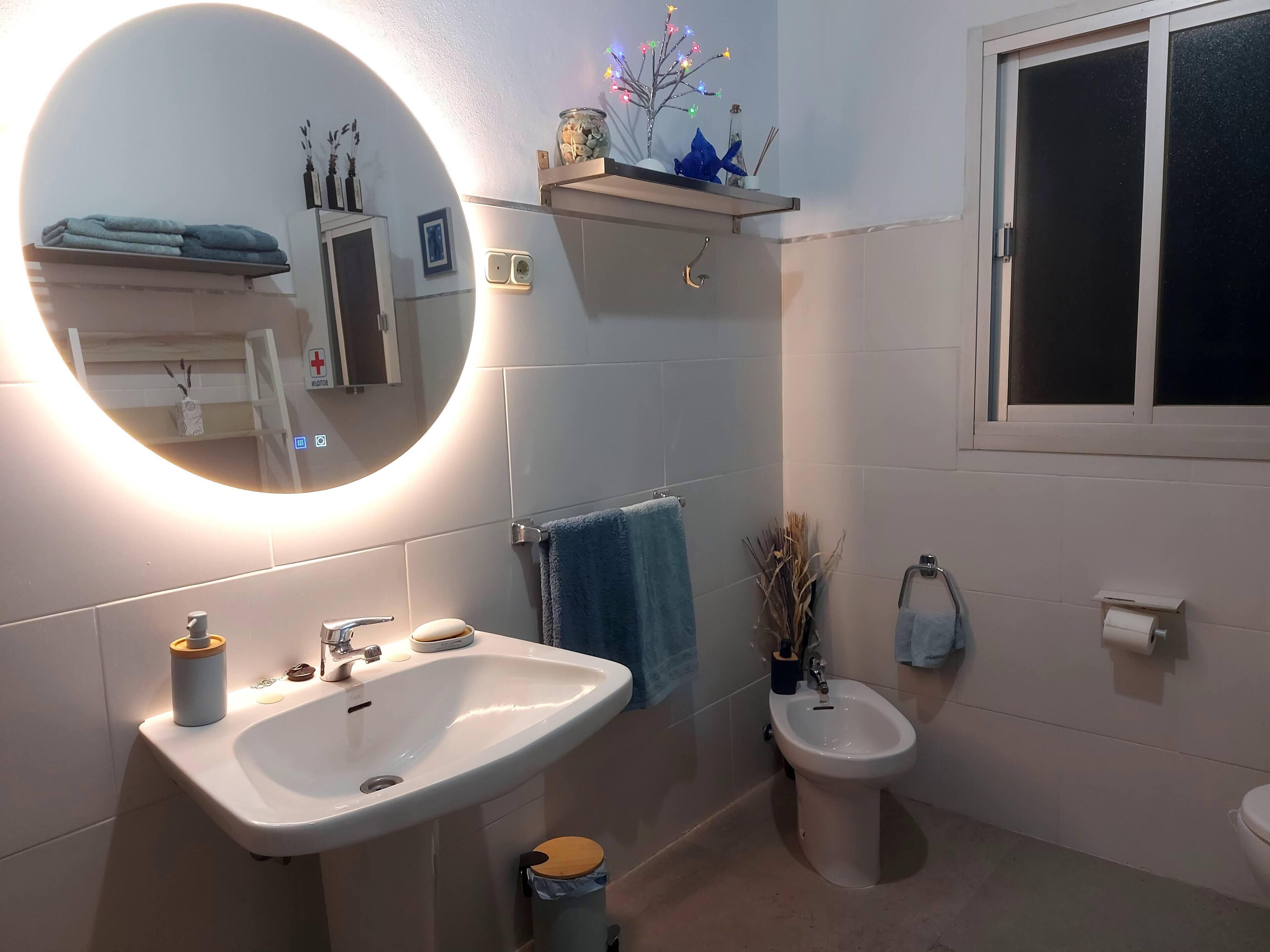Un baño moderno con un espejo grande, un lavamanos, un bidé y un inodoro. Hay un perchero para toallas y una repisa con objetos decorativos.