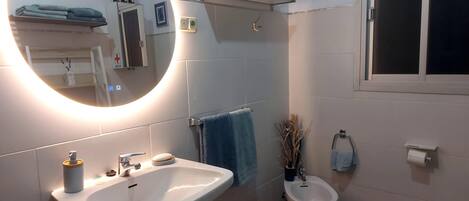 Un baño moderno con un espejo grande, un lavamanos, un bidé y un inodoro. Hay un perchero para toallas y una repisa con objetos decorativos.