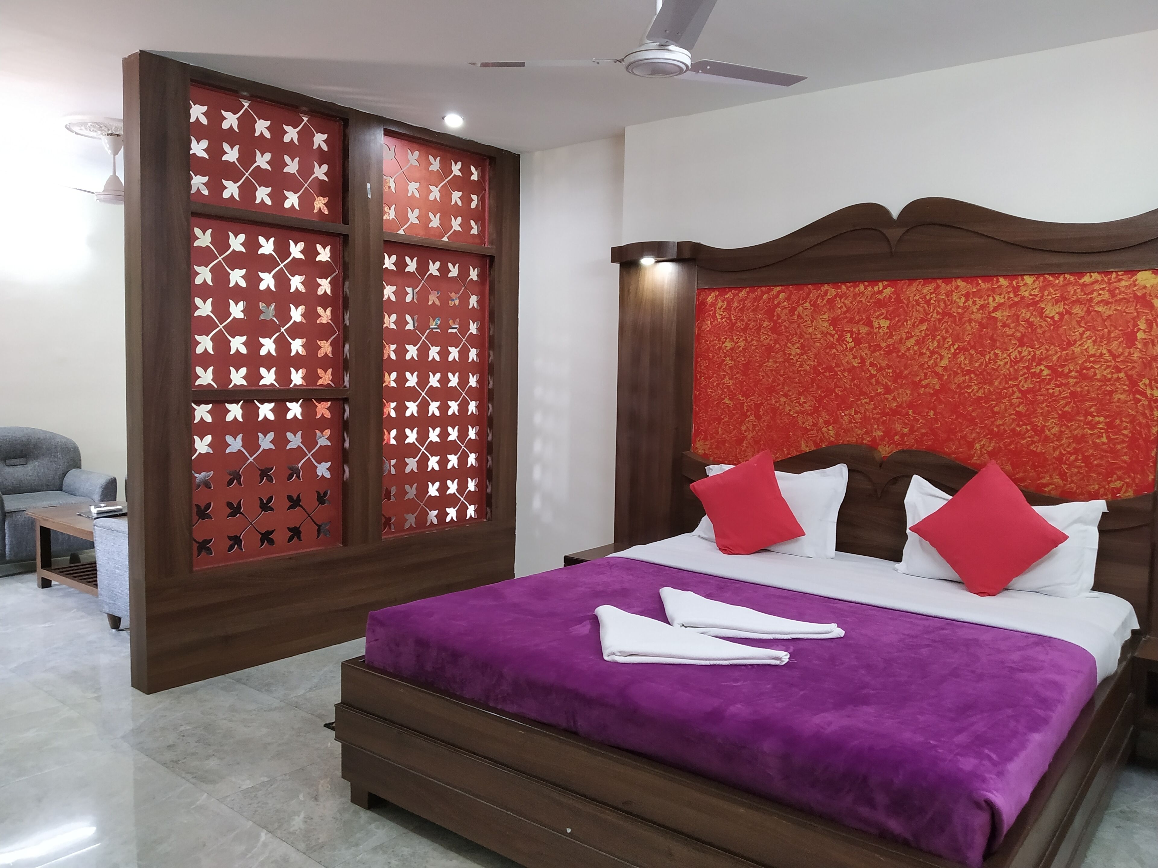 A hotel room with a bed, a wooden headboard, a red patterned partition, and a grey sofa.