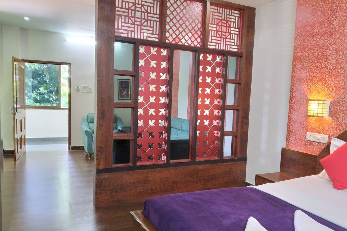 Quarto de hotel com cama, divisória de madeira com painéis decorativos, área de estar com cadeiras e vista para o verde lá fora.