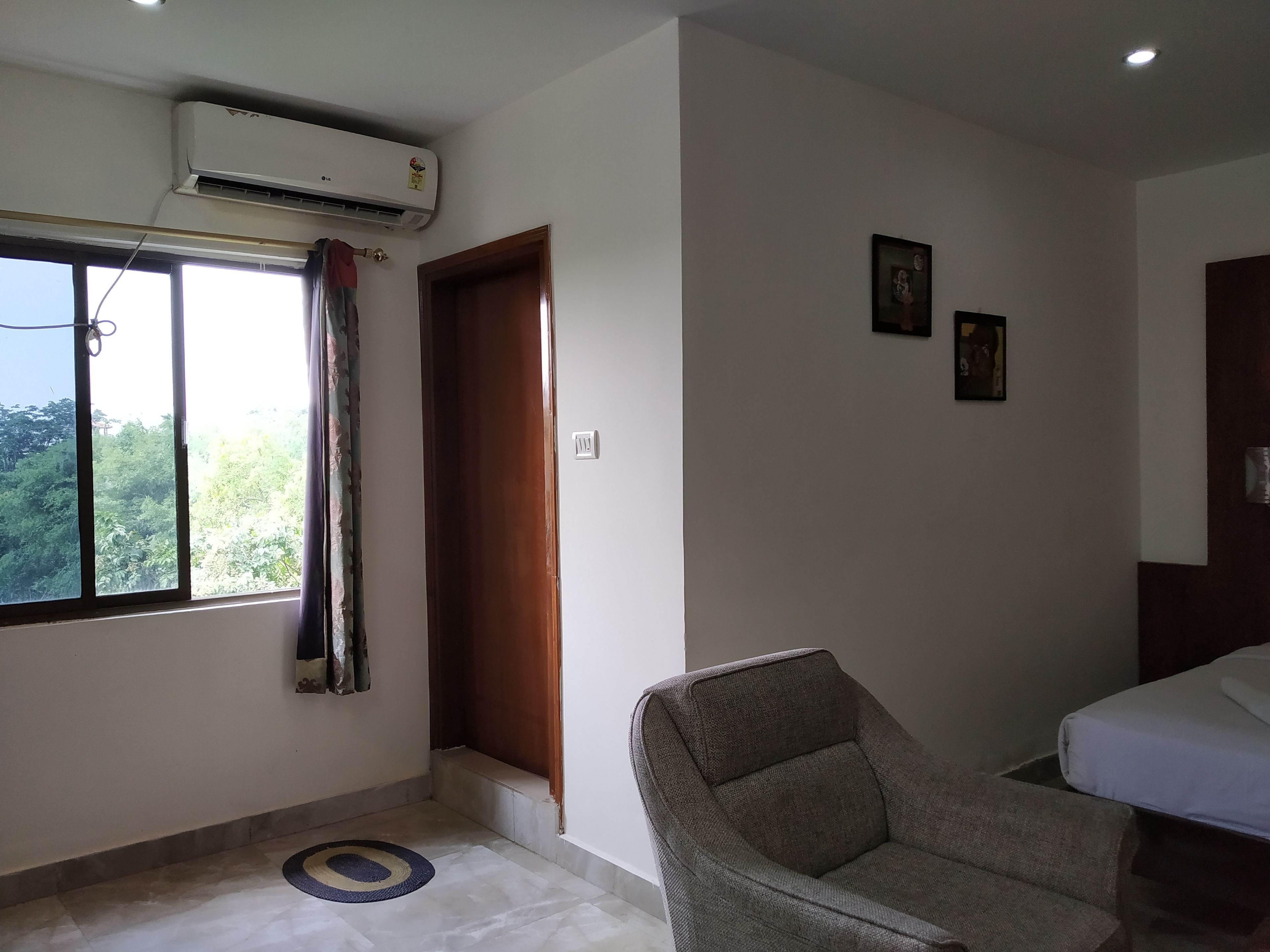 A hotel room with a large window, an air conditioning unit, a wooden door, a grey armchair, and a bed.
