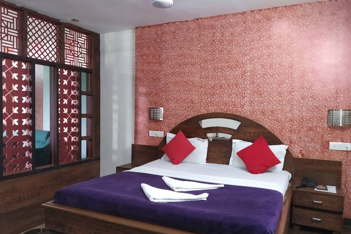 Quarto de hotel com uma cama grande, cabeceira de madeira, almofadas vermelhas e brancas, colcha roxa e mesa de cabeceira com um telefone, além de um painel decorativo na parede.