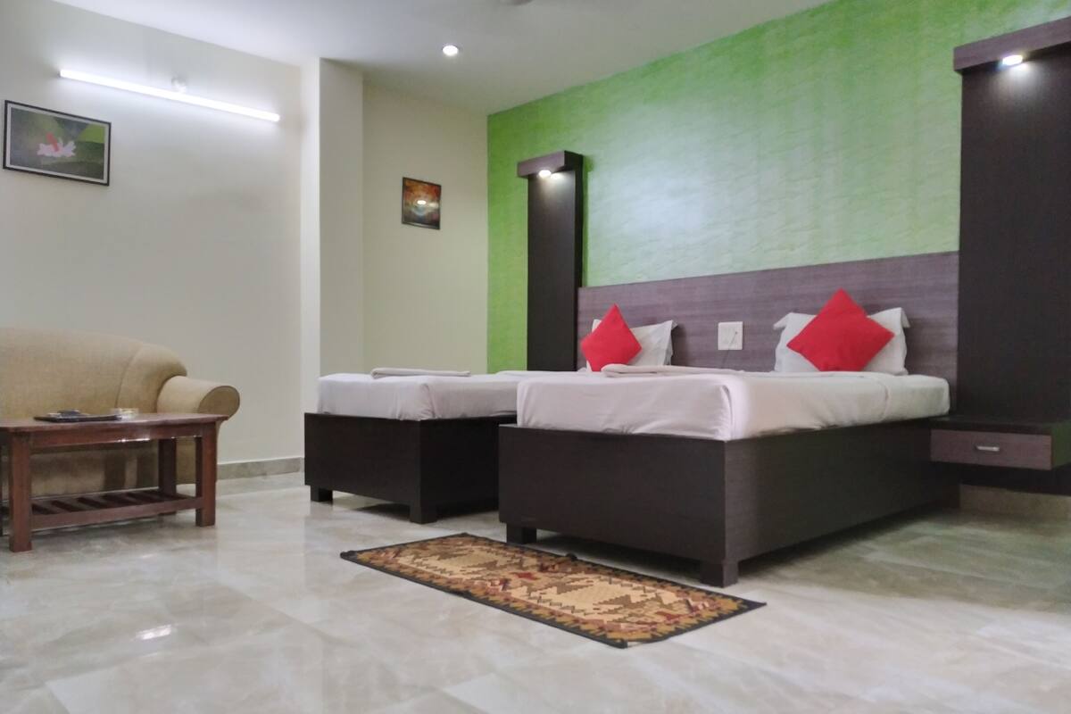 Quarto de hotel com uma cama grande, uma cama menor, um sofá, uma mesa de madeira e uma parede verde decorativa.