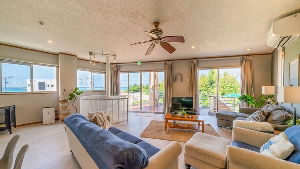 A spacious living room with a large sofa, a coffee table, and a television. There are large windows offering a view of the outdoors. - Lanakai House (Onna)