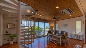 A spacious room with a wooden floor, a dining area with a table and chairs, a spiral staircase, and large windows offering a view of the outdoors. - Lanakai House (Onna)