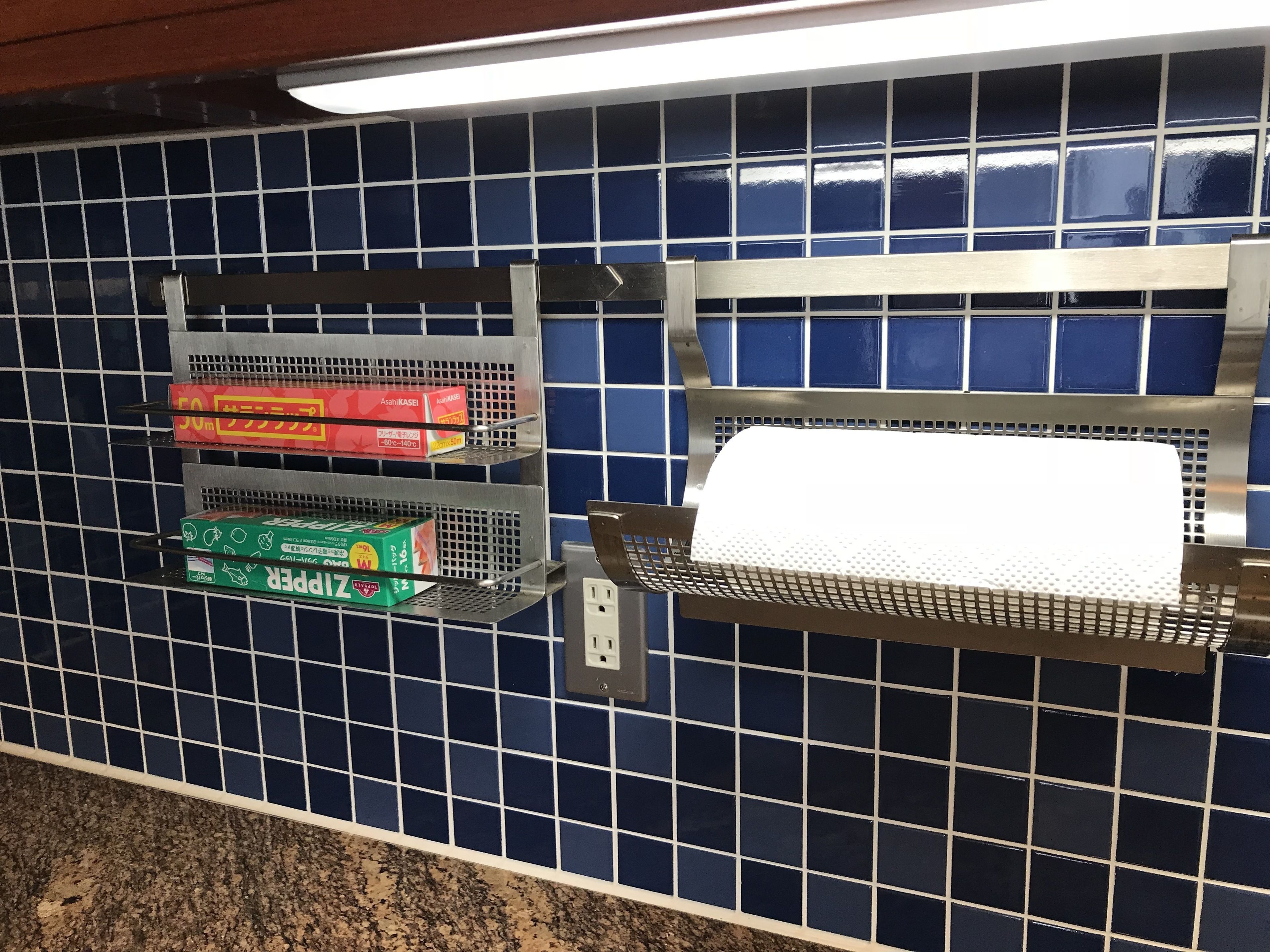 A kitchen with blue tiled backsplash, metal shelves holding kitchen supplies, and a paper towel dispenser.